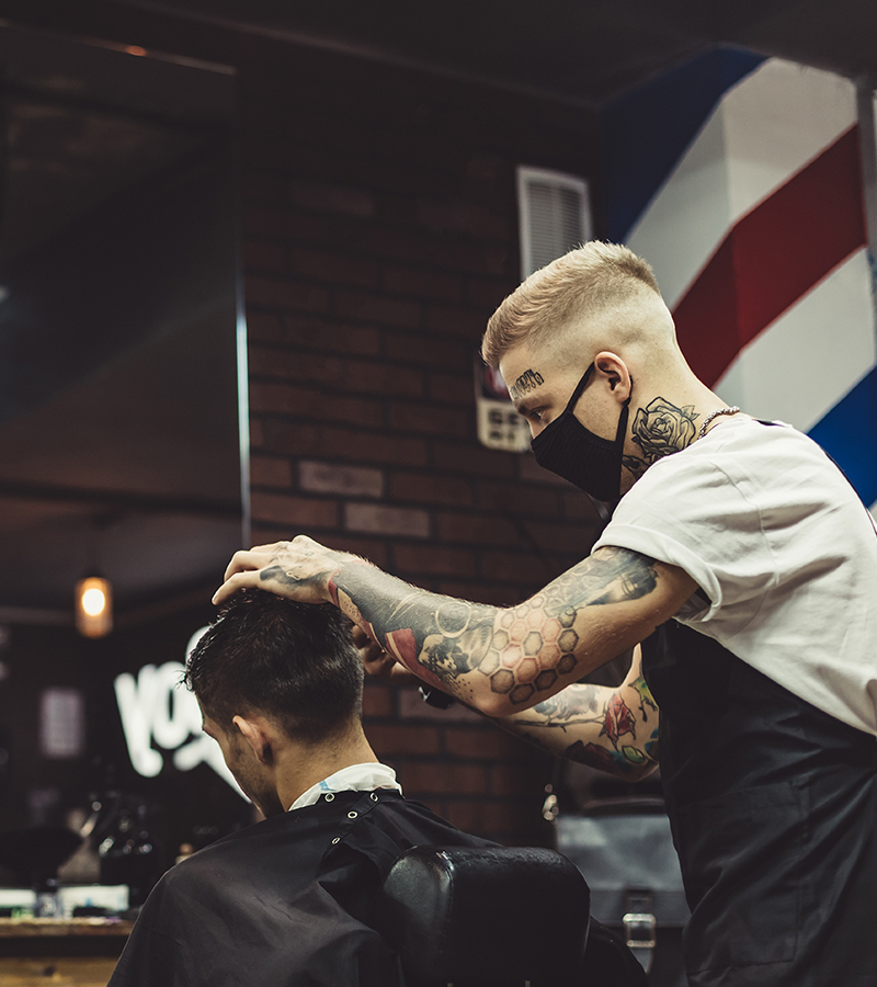 Un barbero, con tatuajes en los brazos y mascarilla negra, recorta el cabello de un cliente. El cliente está sentado de espaldas en la silla de barbería.